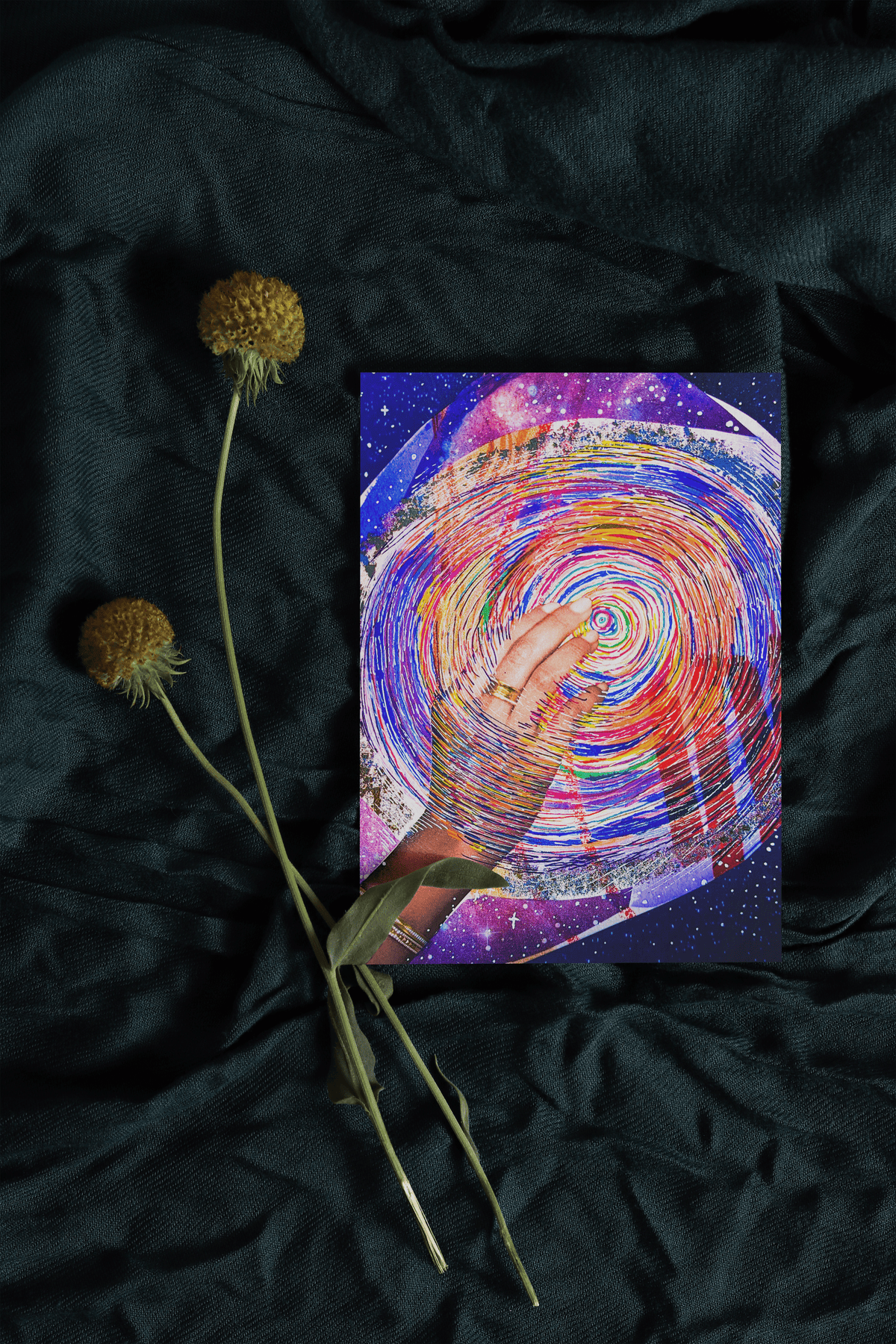 Card sitting on teal fabric background with two dried flowers lying next to it. Card is red and purple concentric line circles with a hand reaching into the center of the circle.