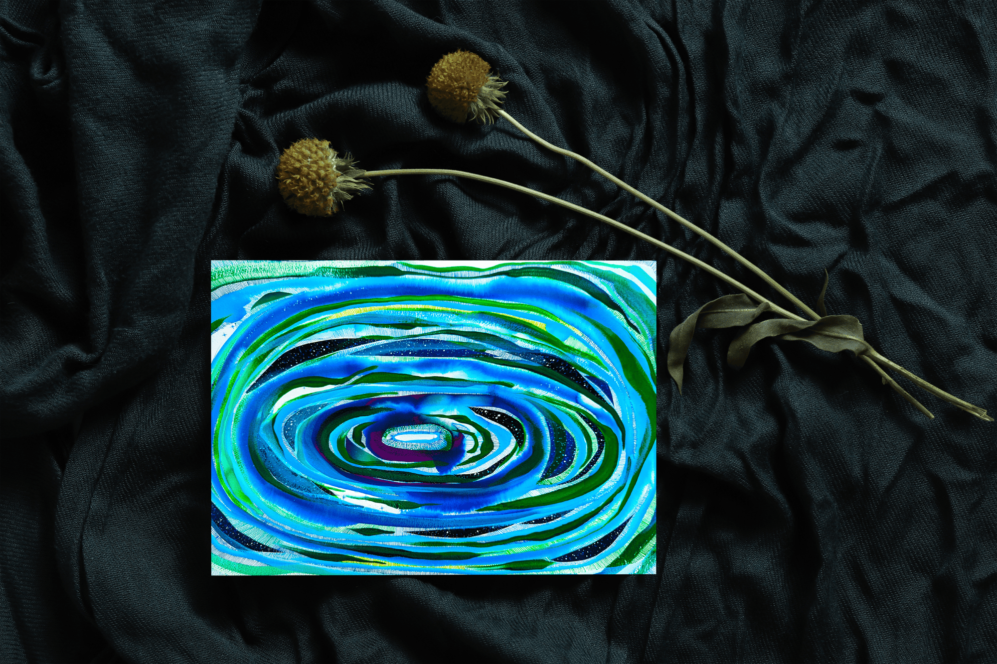 Notecard sitting on teal draped fabric with dried flowers. Card is painting of blue and green concentric ovals 