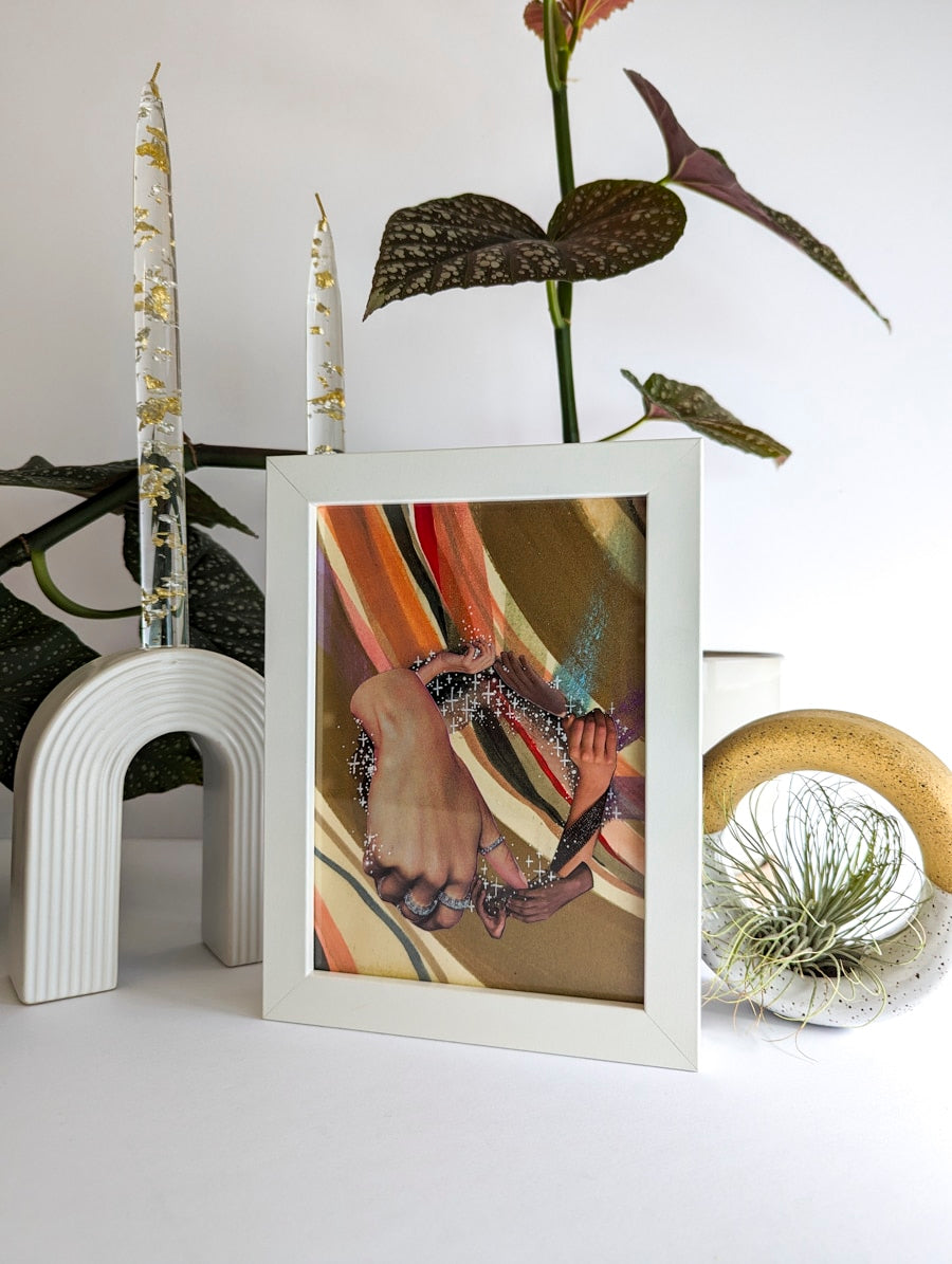 White framed collage on paper. Background is plant and white ceramic candle holder. Art collage shows hands arranged in a circular shape. Multicolor striped background. Bronze and rainbow metallic accents.