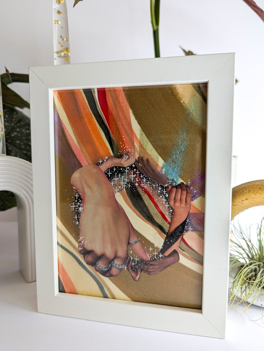 White framed collage on paper. Background is plant and white ceramic candle holder. Art collage shows hands arranged in a circular shape. Multicolor striped background. Bronze and rainbow metallic accents.