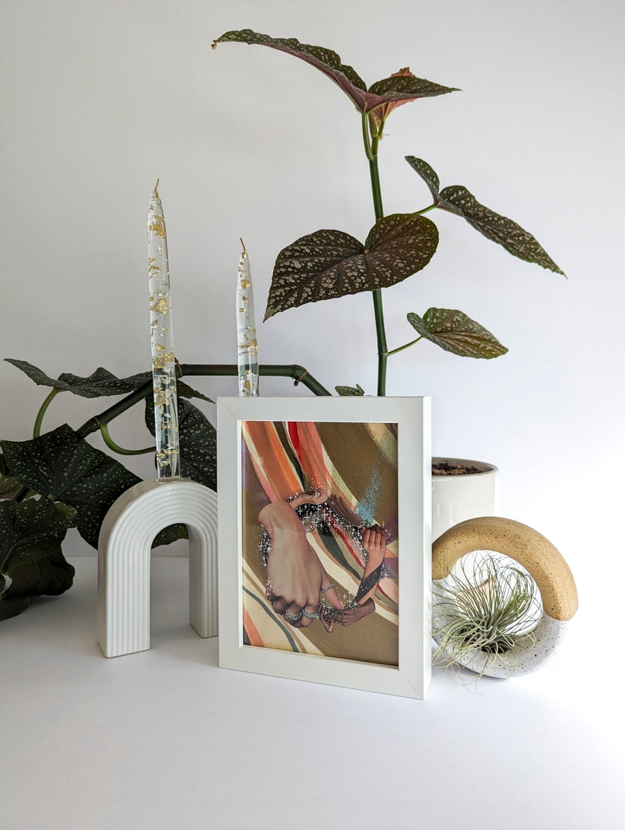 White framed collage on paper. Background is plant and white ceramic candle holder. Art collage shows hands arranged in a circular shape. Multicolor striped background. Bronze and rainbow metallic accents.