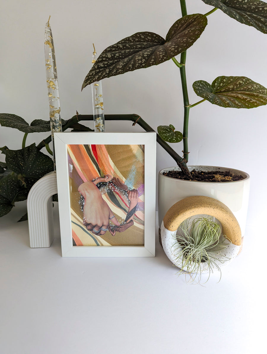 White framed collage on paper. Background is plant and white ceramic candle holder. Art collage shows hands arranged in a circular shape. Multicolor striped background. Bronze and rainbow metallic accents.