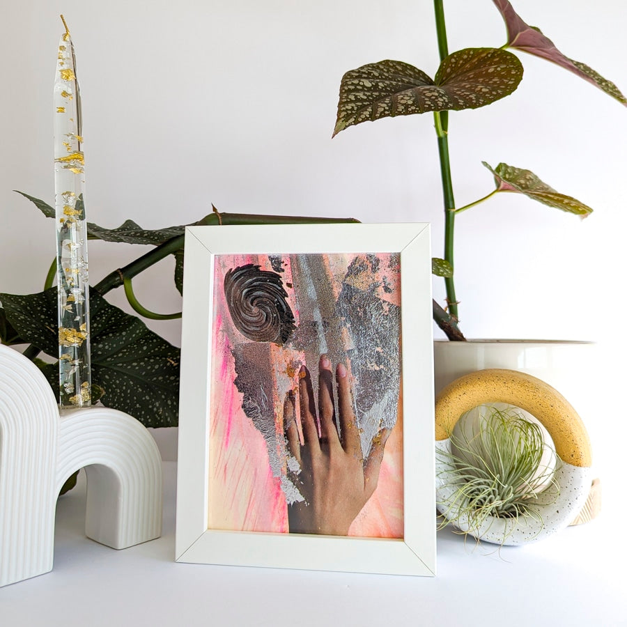 White framed collage on paper. Background is plant and white ceramic candle holder. Art collage shows a hand on pink background with a swirling galaxy shape and silver metallic accents.