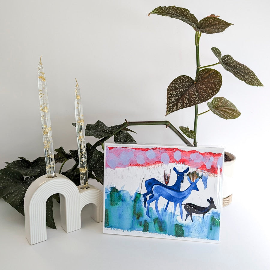 Art print on paper against background with plant and white ceramic candle holder. Art print shows a group of blue deer painted in watercolor with a red and lavendar sky in background. 