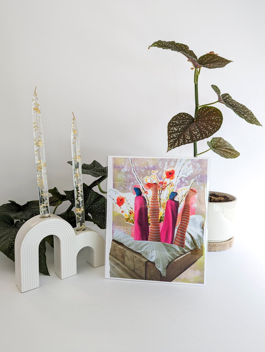 Art print on paper against background with plant and white ceramic candle holder. Art print is a collage of a bed with 4 legs sticking straight up out of it, one set of legs is wearing bright pink pants and the other orange and white striped tights. Orange flowers and twigs layered on lavender in background.