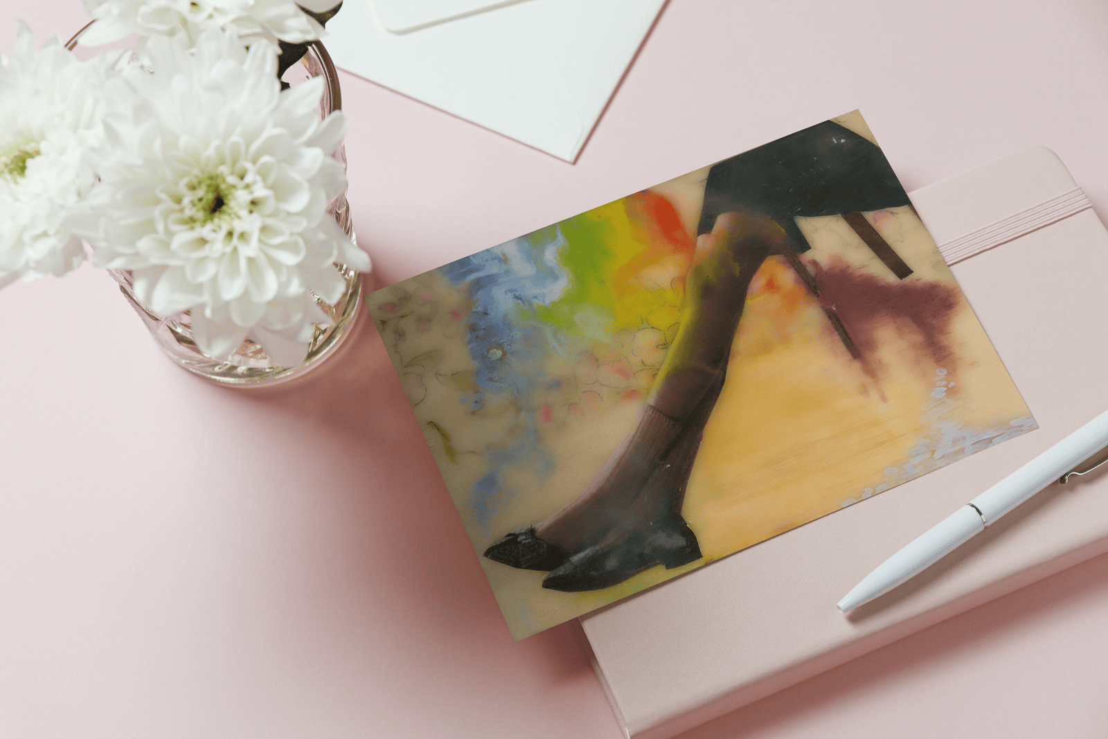 Card with a woman's legs wearing black shoes, pink socks and a black skirt stretched out sitting on a chair. Background of card shows wood grain, a dissolved rainbow Blue and flower patterned fabric bits. Card sits on a light pink table with a white pen, pink journal and white flower in a vase next to it.