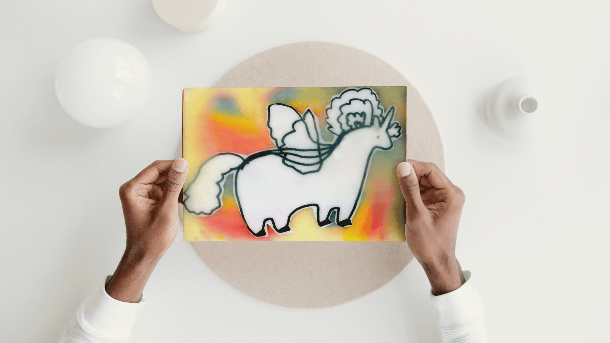 Art print of black and white chubby pegacorn on rainbow background being held by two hands over a white table 