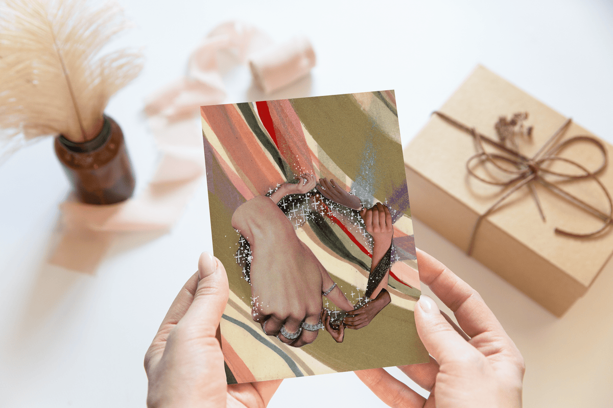 Two hands holding a card, the card shows a circle of hands holding each other. Light brown decor objects in background of photo 