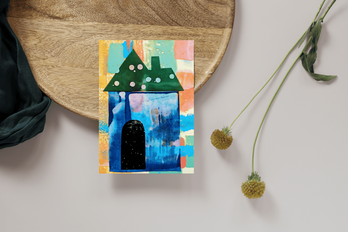 Card with simple house painting. House is blue with a starry sky for a door. Colorful background on card. Sitting on a wood circle with teal fabric draped next to it and two dried flowers.